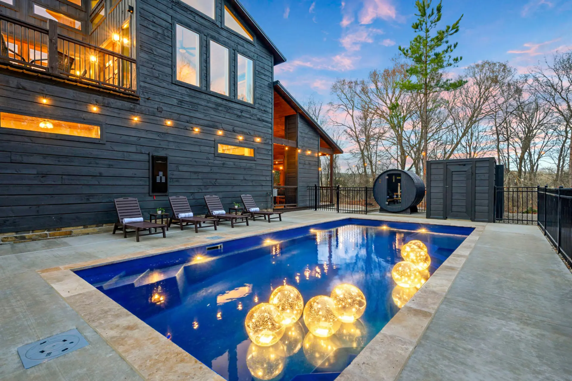 This backyard is absolutely stunning with a heated pool, lounge chairs, a sauna pod, and views of the woods at sunset.
