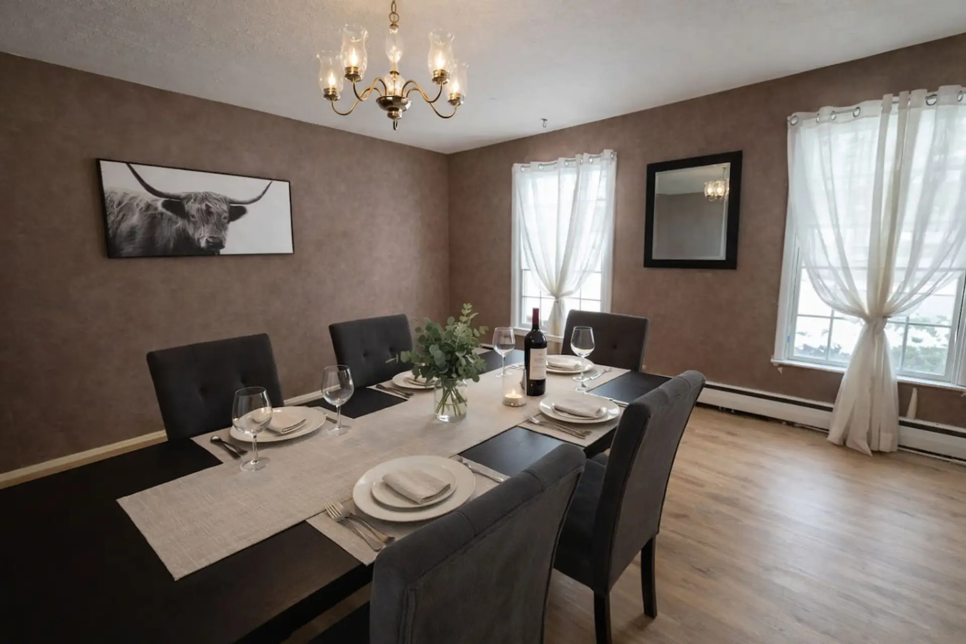 This sophisticated dining room features a large table with seating for six, perfectly arranged under the warm glow of a classic chandelier for an elegant mealtime experience. Earthy-toned walls and rustic-chic decor create an intimate and refined atmosphe