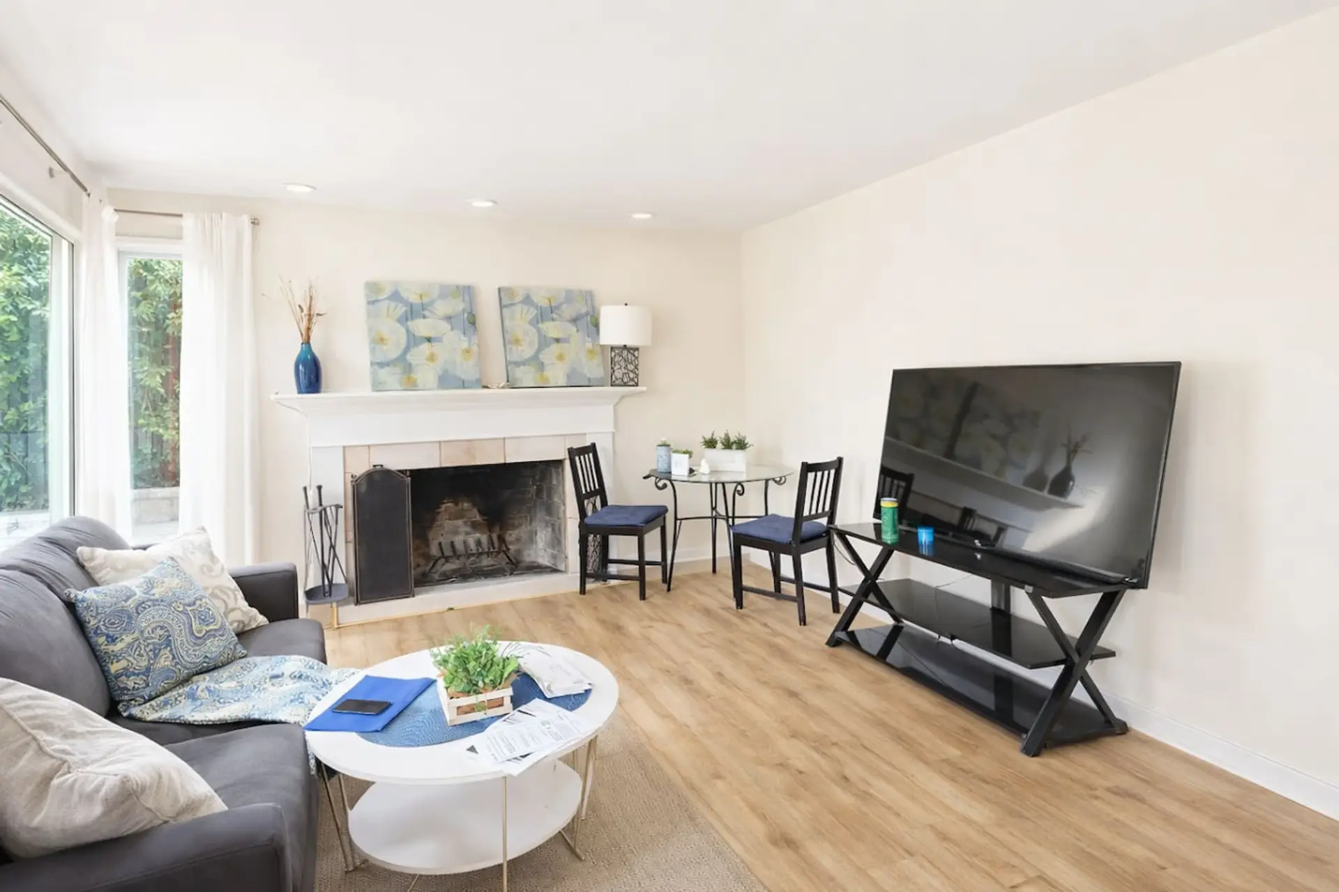 This inviting living area features a charming fireplace and a large flat-screen TV, creating a cozy and functional space for relaxation or movie nights. Modern wood flooring and neutral tones are complemented by stylish blue accents and a secondary bistro