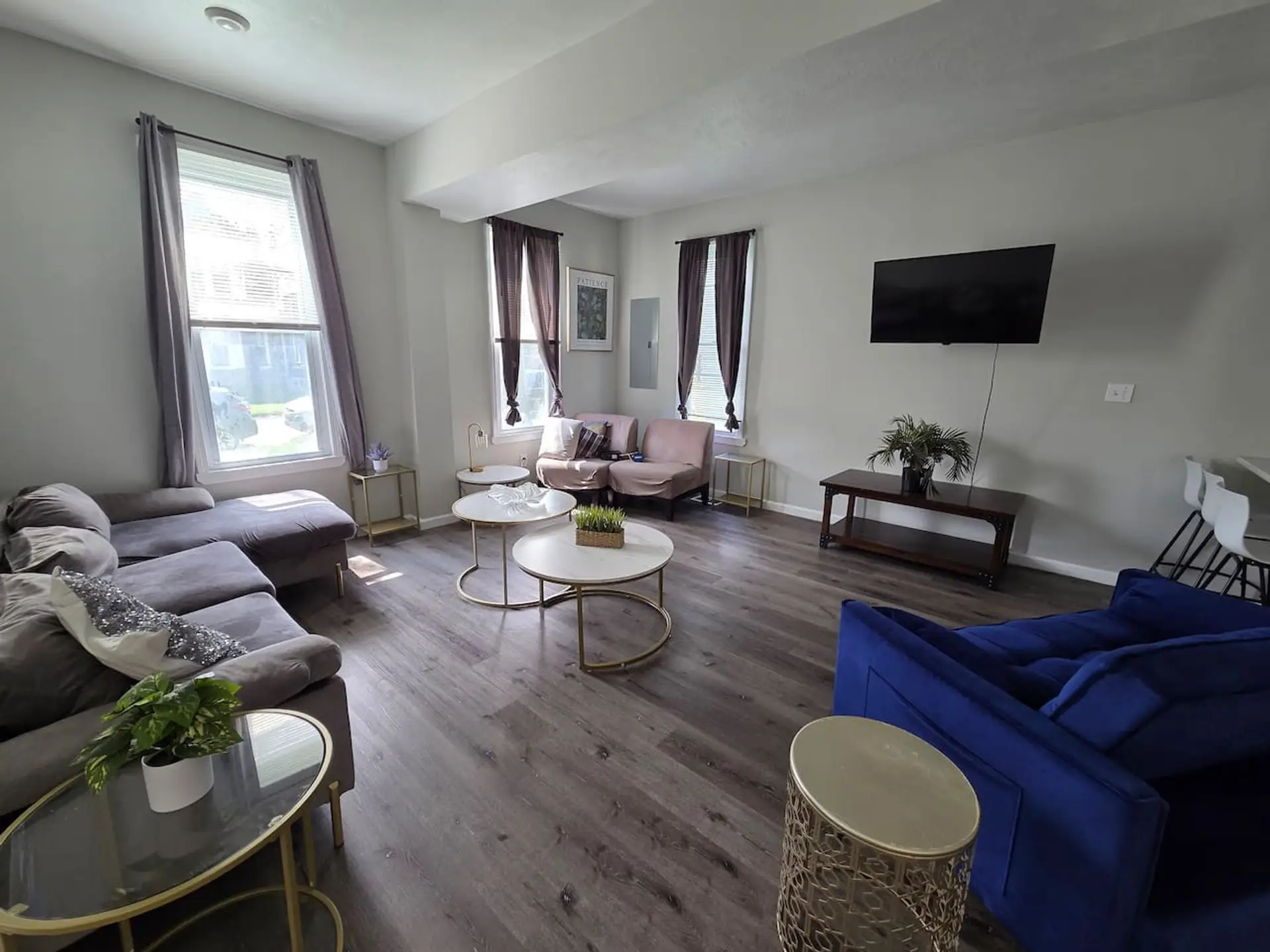 This bright and airy open-concept living area seamlessly connects to the kitchen, featuring sleek grey wood-toned flooring and modern recessed lighting throughout. The space offers ample seating with a plush velvet sectional and a striking cobalt blue lov