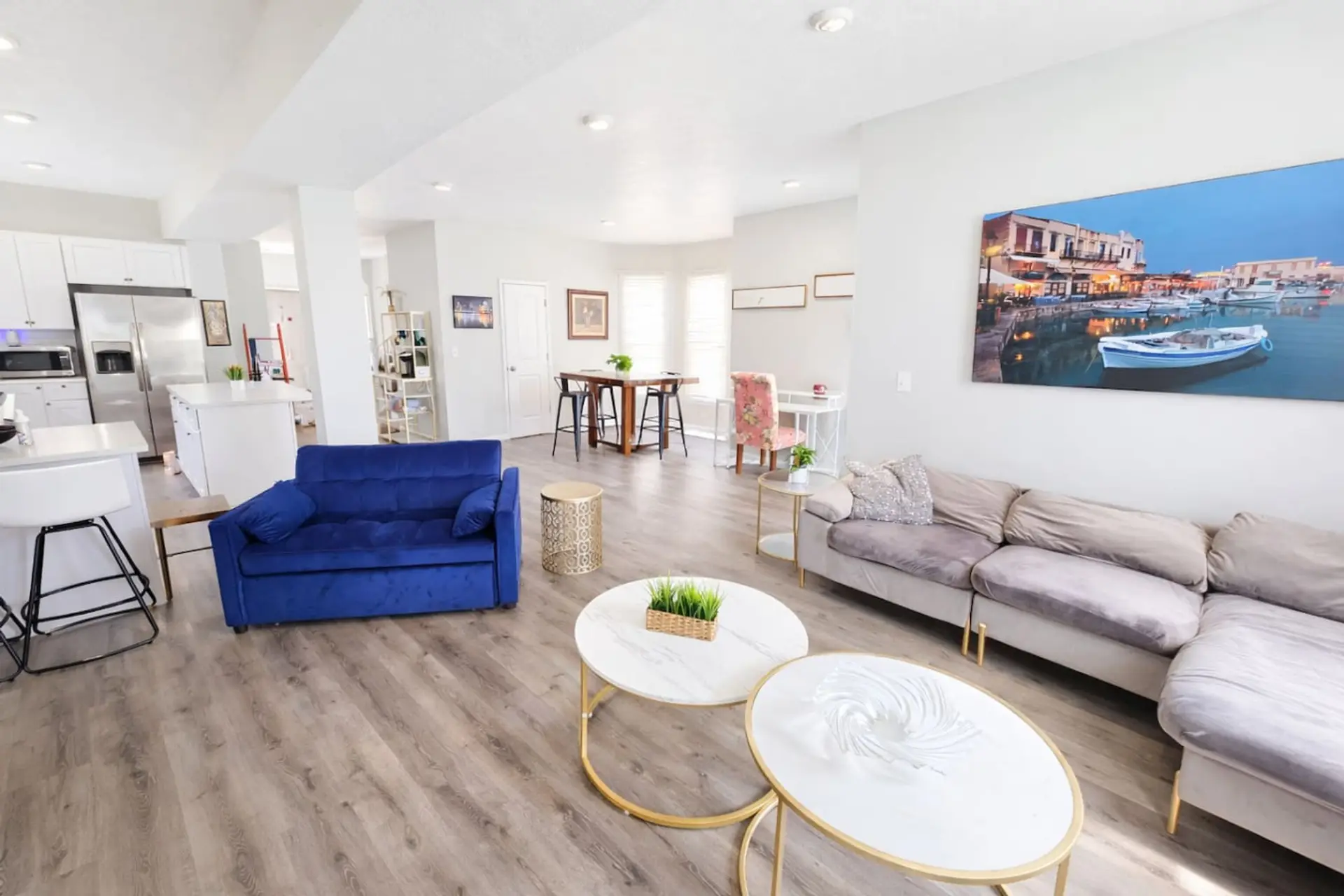 This bright and airy open-concept living area seamlessly connects to the kitchen, featuring sleek grey wood-toned flooring and modern recessed lighting throughout. The space offers ample seating with a plush velvet sectional and a striking cobalt blue lov