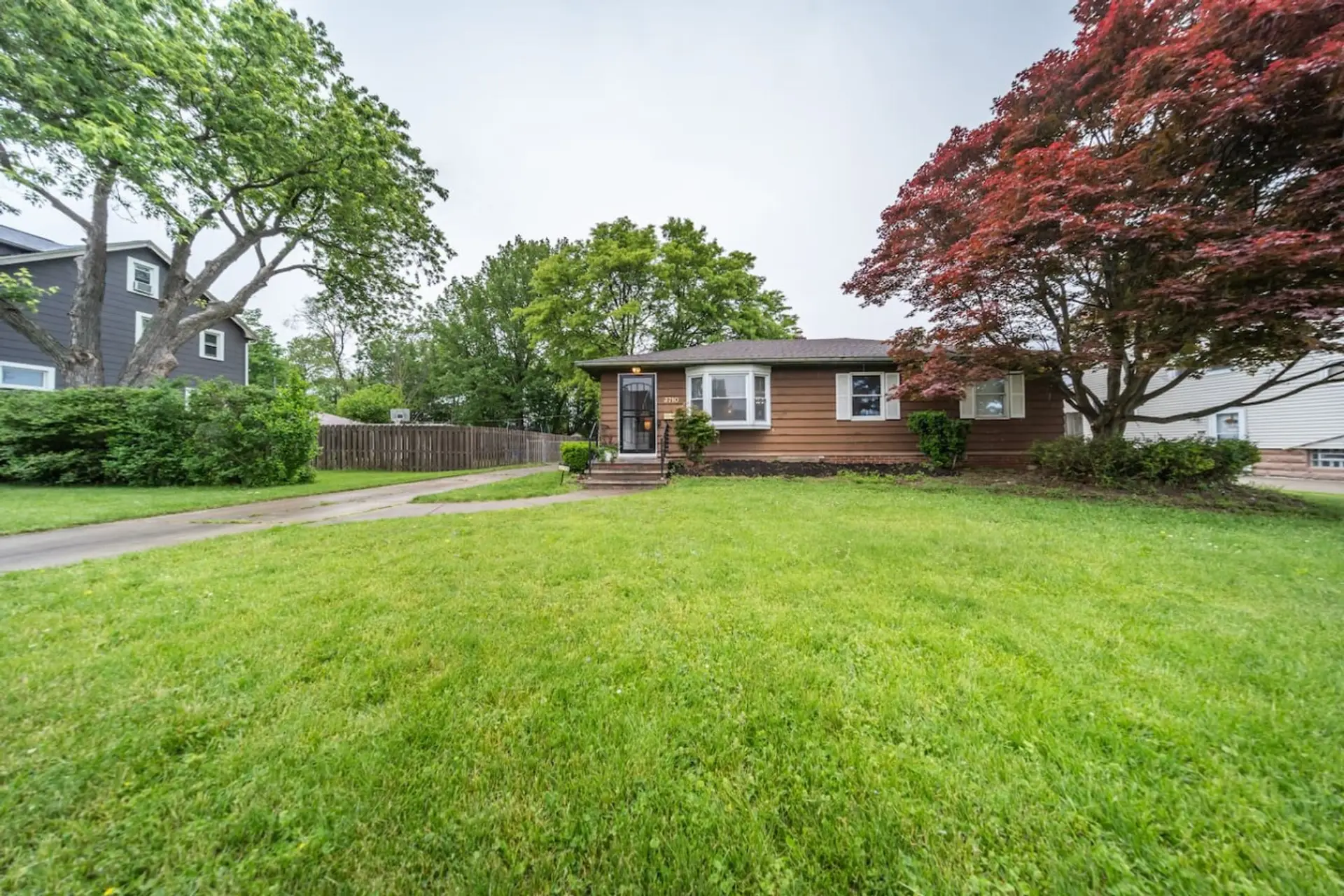 This charming home features warm, wood-toned siding that blends beautifully with the surrounding greenery, set perfectly in the center of a sprawling, vibrant lawn and shaded by mature, leafy trees.