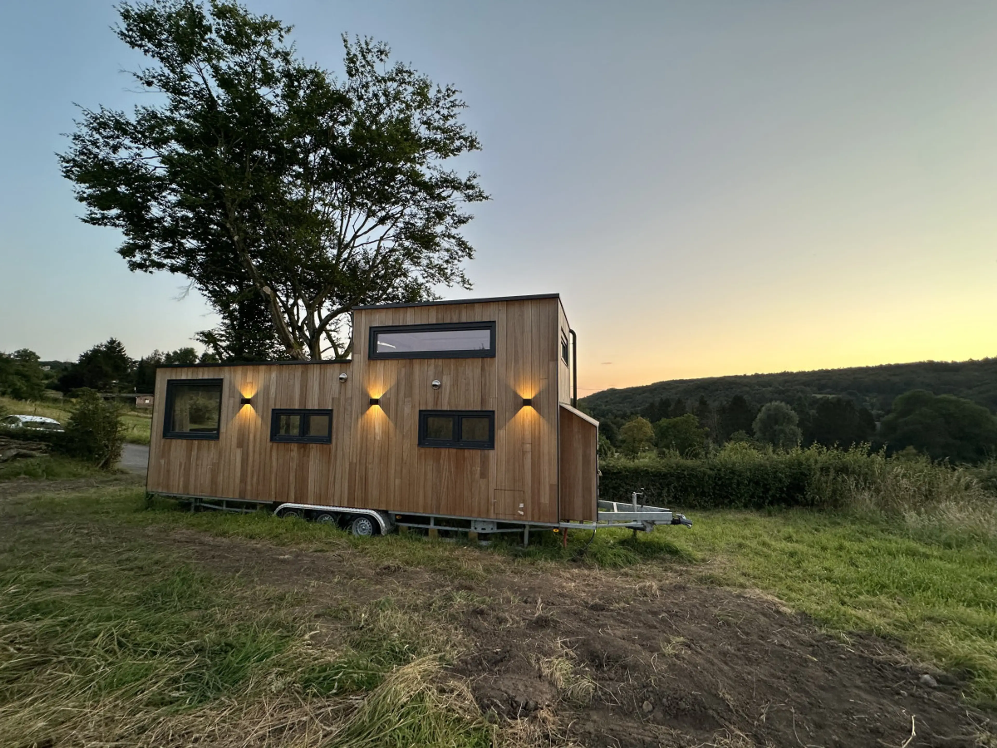 Face arrière d'une tiny house éclairée avec vue sur coucher de soleil, fenêtres panoramiques à l'étage offrant une vue à 360°