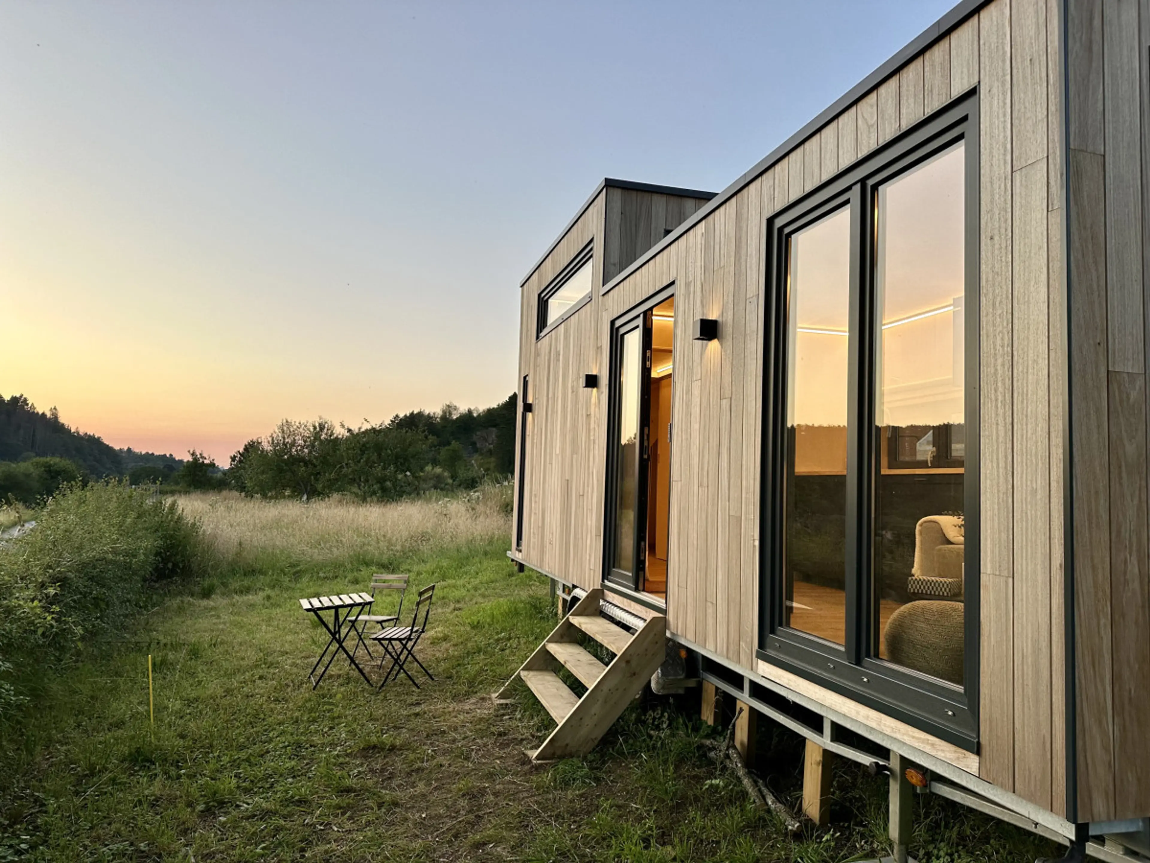 Tiny house avec façade avant vitrée face à un coucher de soleil, petit escalier d'accès et table extérieure pour apéro en plein air.