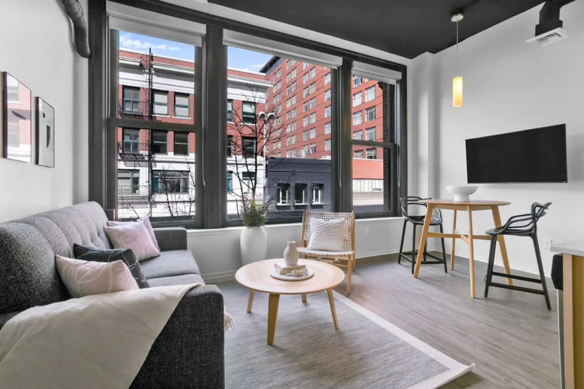 A sectional sofa and round wood coffee table sit beside large windows overlooking a brick building.