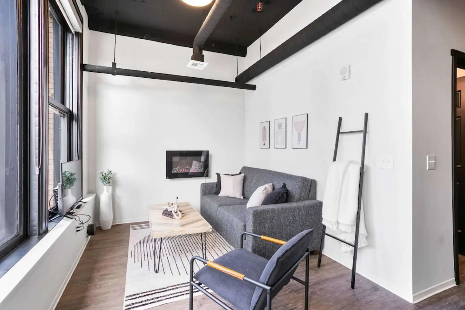 A gray sofa and armchair sit on a striped rug below exposed black ceiling beams, with framed art on the white brick wall.