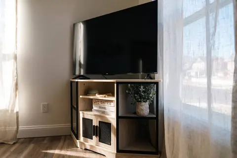 TV on a wooden stand beside a window with sheer curtains