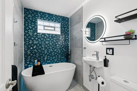 Bathroom with a freestanding tub, teal tile, a round lit mirror, and a small sink