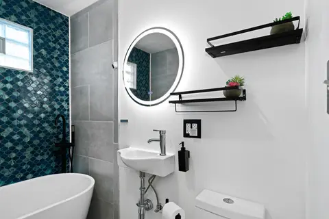 Small sink below a round lit mirror and black floating shelves