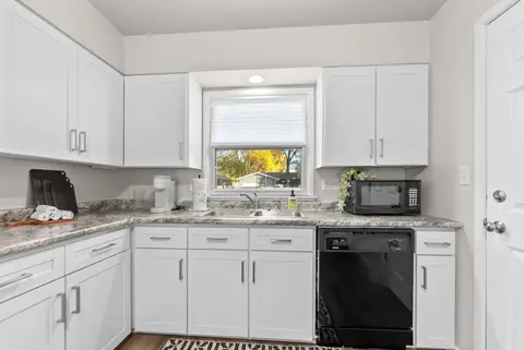 Kitchen sink under a window, between white cabinets and a black dishwasher