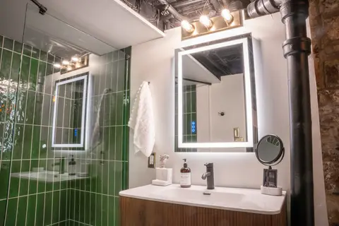 Wood vanity under a backlit mirror, beside the green-tiled shower