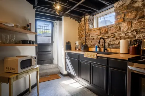 Kitchen with black cabinets and butcher block counters below a stone wall, beside the entry door and steps