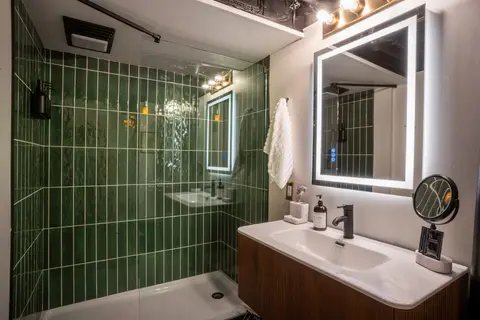 Bathroom with a green-tiled glass shower, a backlit mirror, and a wood vanity