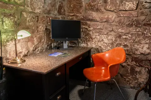 Desk with a monitor, green banker’s lamp, and orange chair against the stone wall