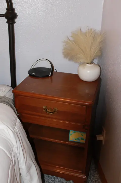 Wooden nightstand with a vase of dried grass beside the bed