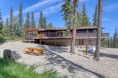 Back of the house with a long deck, a pergola, and a picnic table among the pines