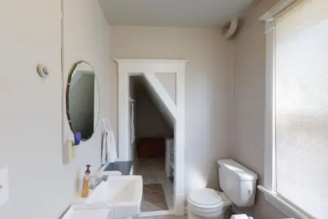 Bathroom with a pedestal sink, a round mirror, and a toilet beside a shaded window