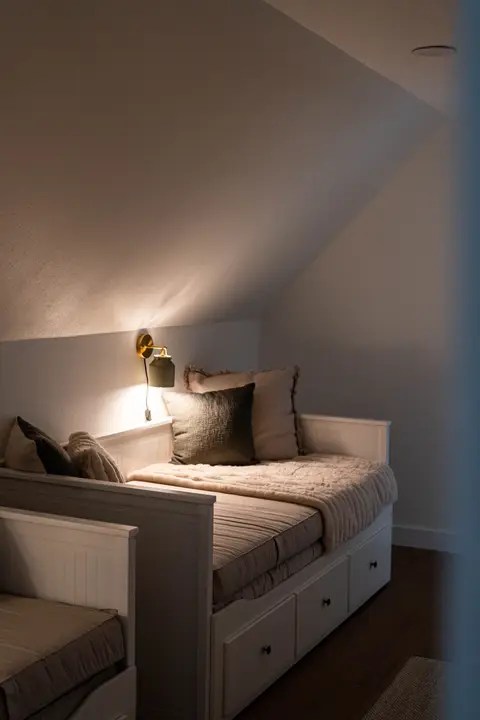 Twin daybed beneath the sloped ceiling with a warm reading light and soft bedding