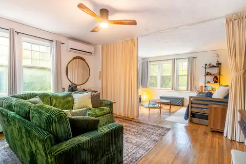 Living room with a green velvet sectional, a ceiling fan, and a mini-split, with the sleeping alcove beyond the curtain