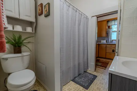 Bathroom with a toilet, open shelves, and a tub behind a shower curtain, with the kitchen through the door