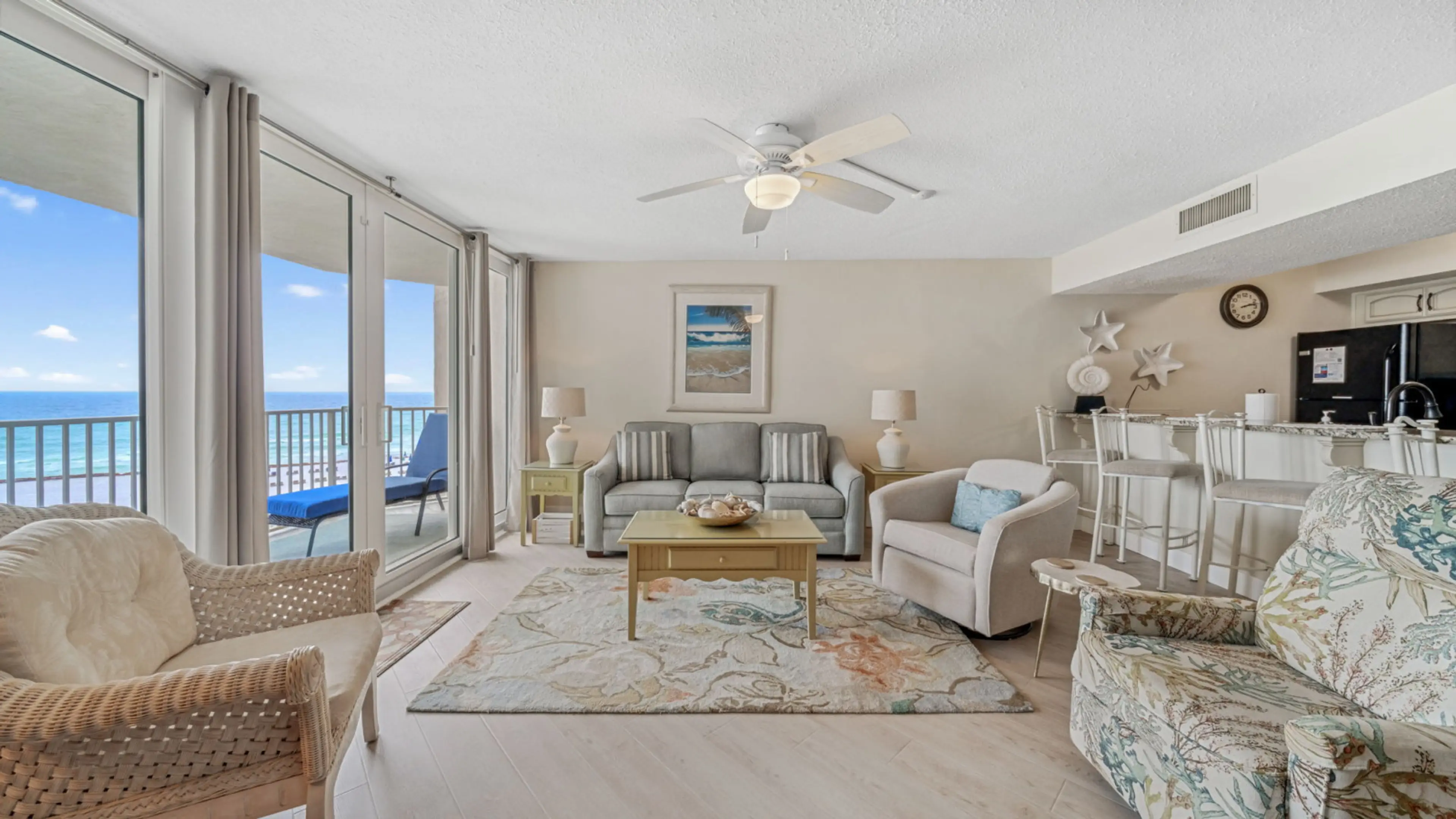 This inviting living room offers a coastal retreat with a breathtaking Gulf-front view. The soft neutral palette is accented by ocean-inspired decor, including shell and starfish wall art. Floor-to-ceiling glass doors flood the space with natural light, l
