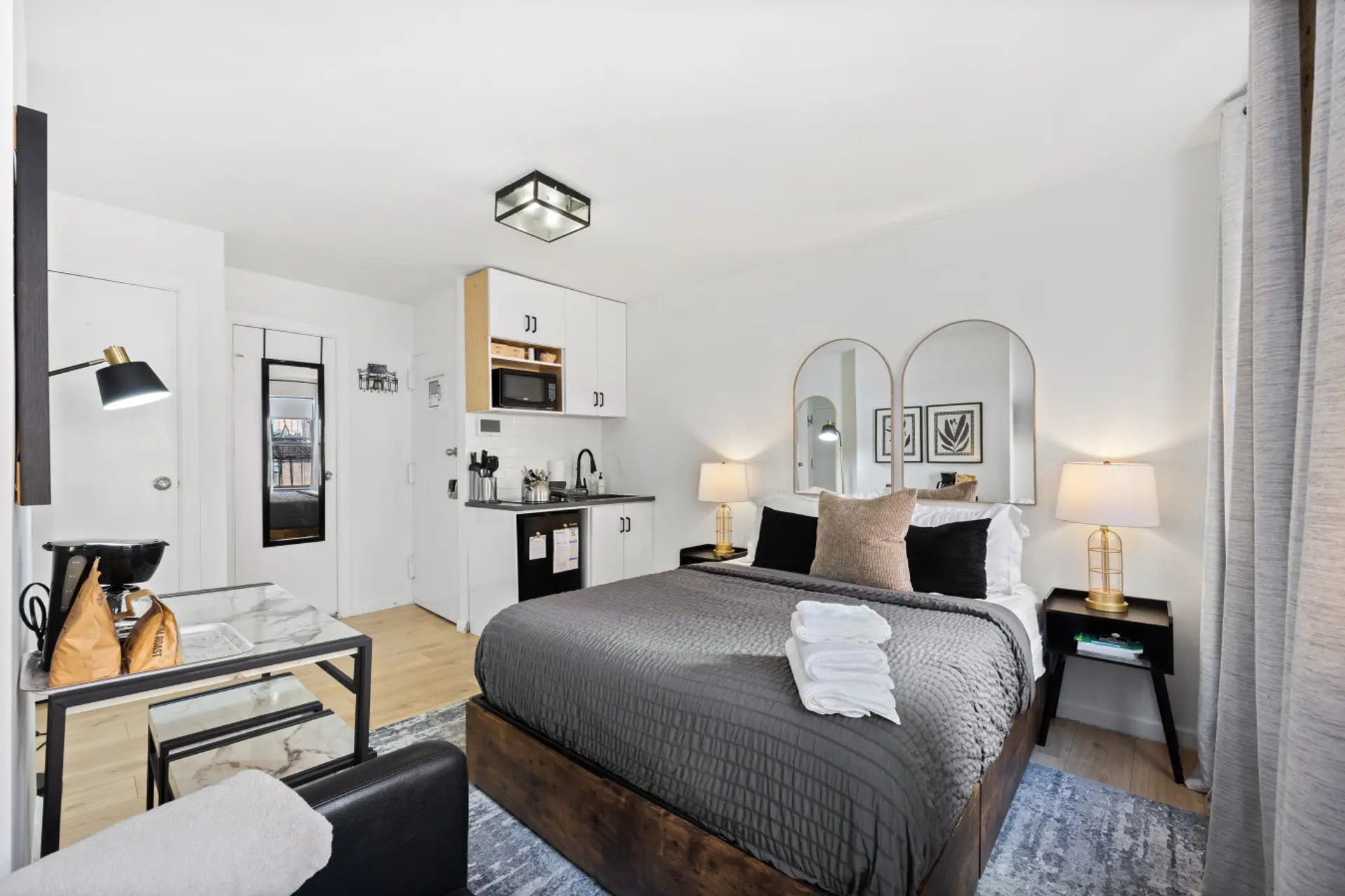 Well-lit room with white bedding, gray pillows, nightstands, kitchenette, table, chair, coffee maker, and wall mirrors.