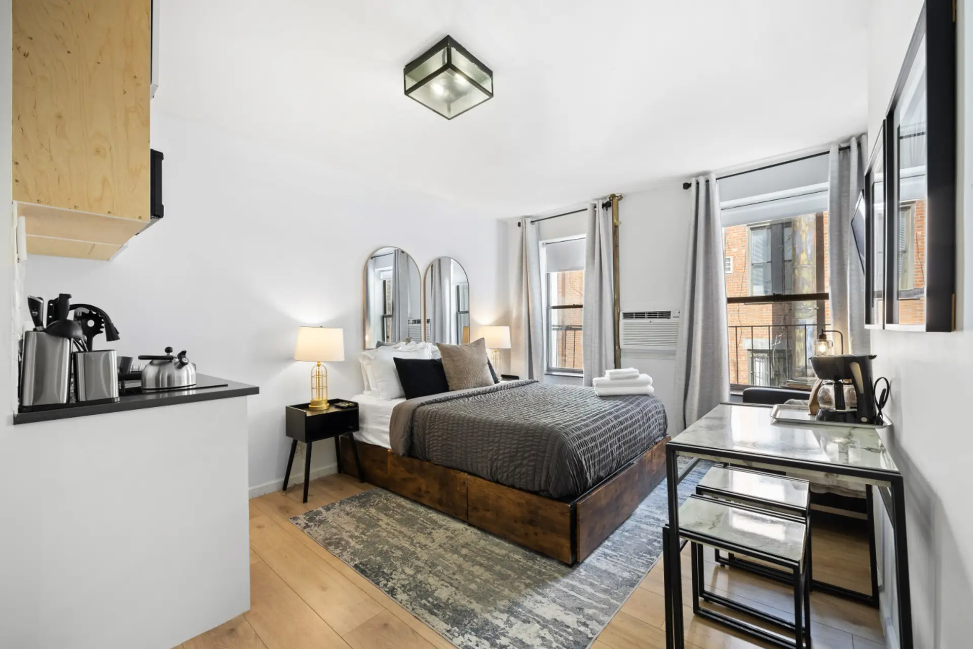 Cozy bedroom with a dark wooden bed, gray bedding, nightstands with lamps, a dining table, coffee maker, and white cabinets. Window with curtains and AC unit.