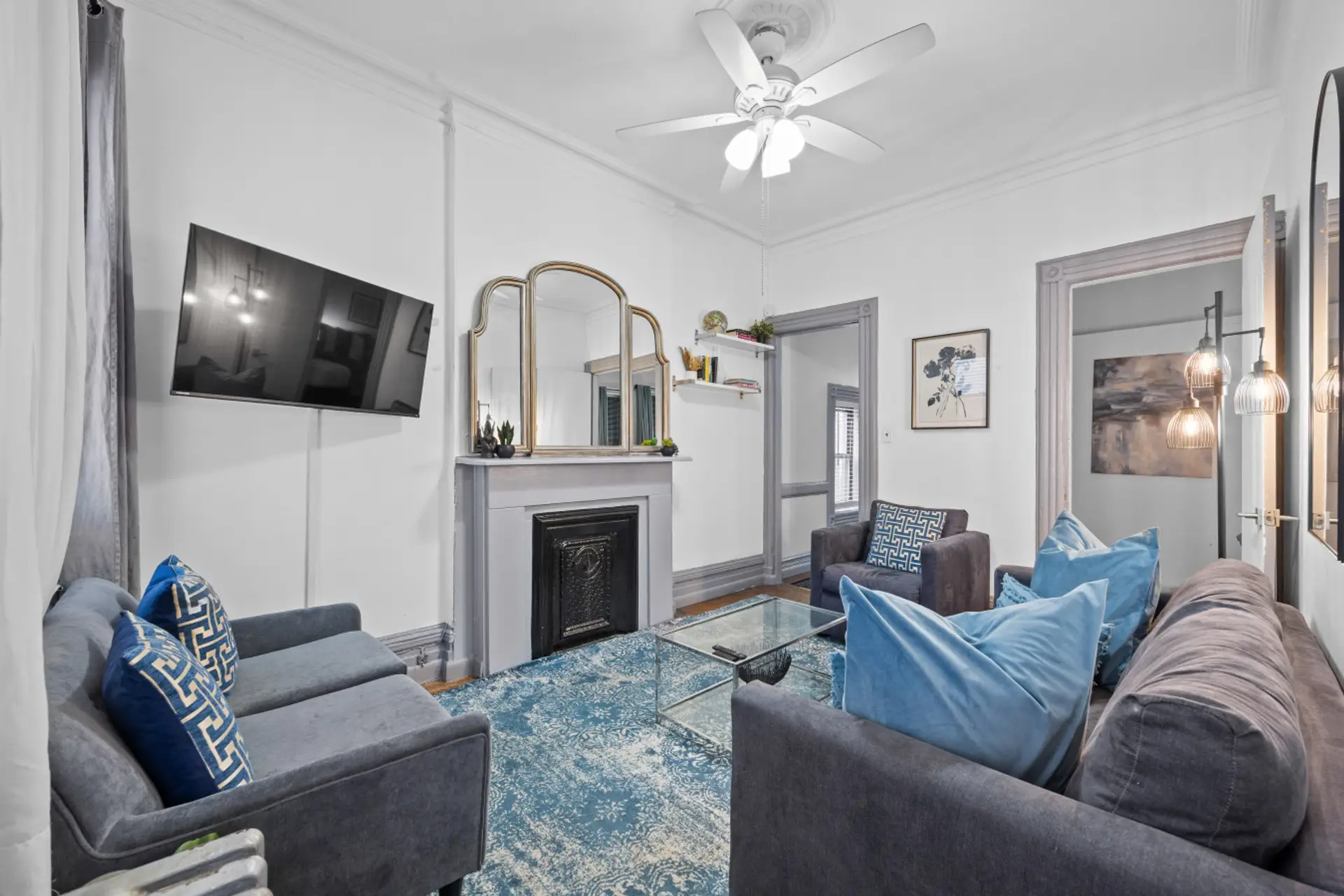 This inviting living room features two comfortable sofas, a cozy armchair, a flat-screen TV, and a decorative fireplace. A stylish mirror, a glass coffee table, and a blue patterned rug complete the elegant space.