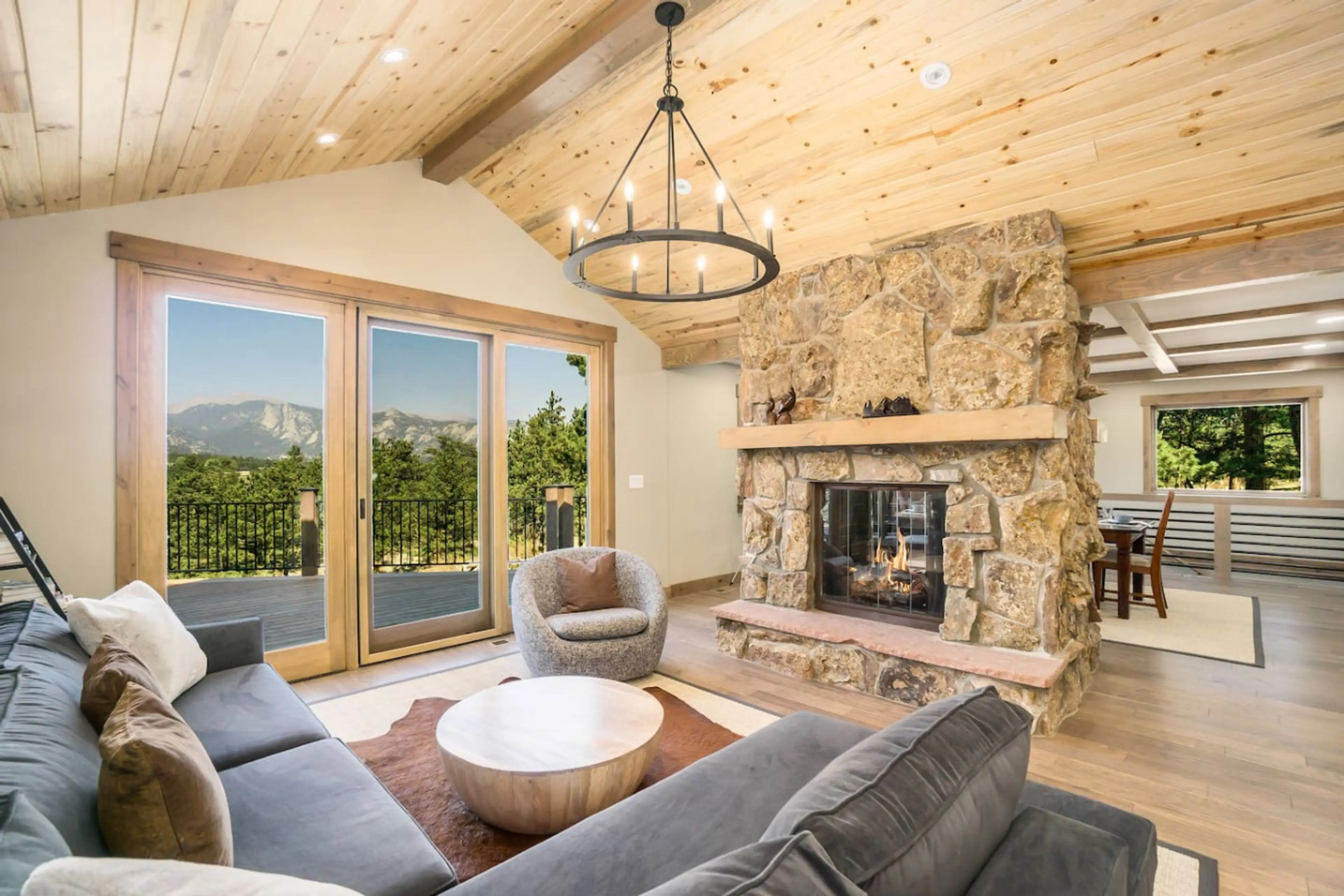 Open-concept living room with two-sided stone gas fireplace and vaulted wood ceilings — a luxury mountain home interior with framed mountain views.