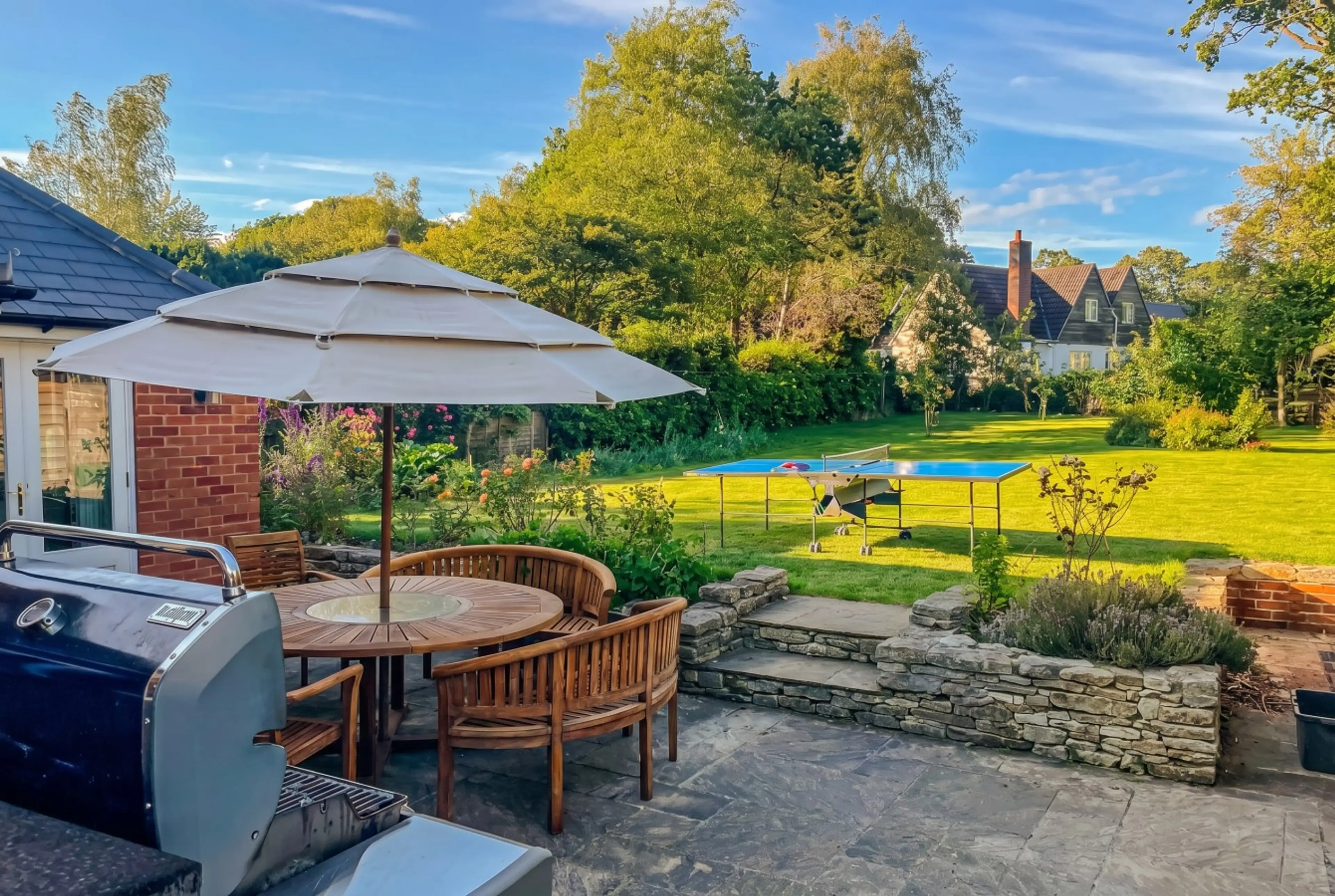 Golden-hour patio with BBQ, teak dining set, and table tennis beyond.