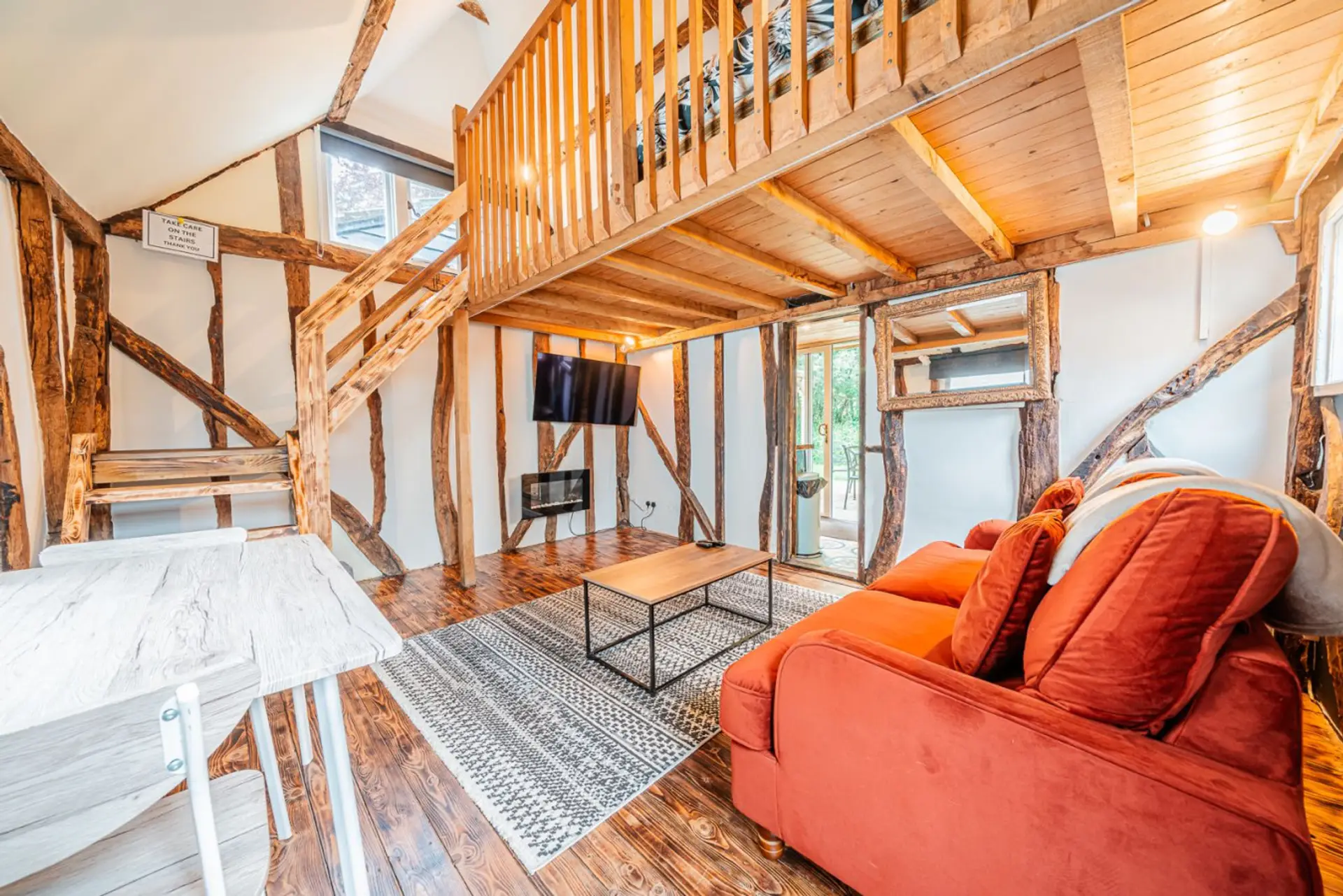 A place to unwind indoors, with an orange sofa, coffee table, and wall-mounted TV beneath a vaulted timber-framed ceiling with a mezzanine level above.