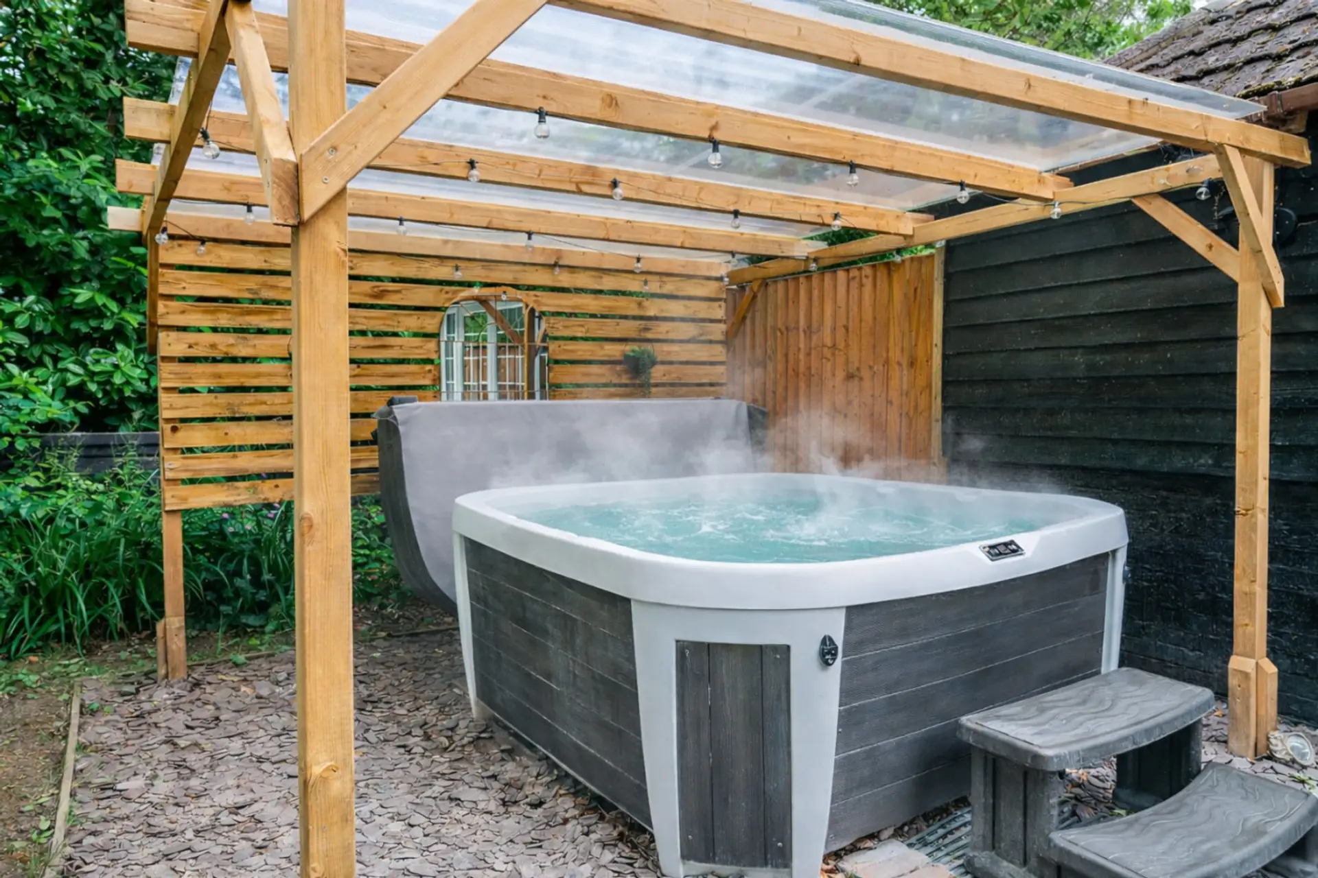 A steaming hot tub sits beneath a timber pergola with a clear polycarbonate roof, set within a fenced enclosure surrounded by mature planting.