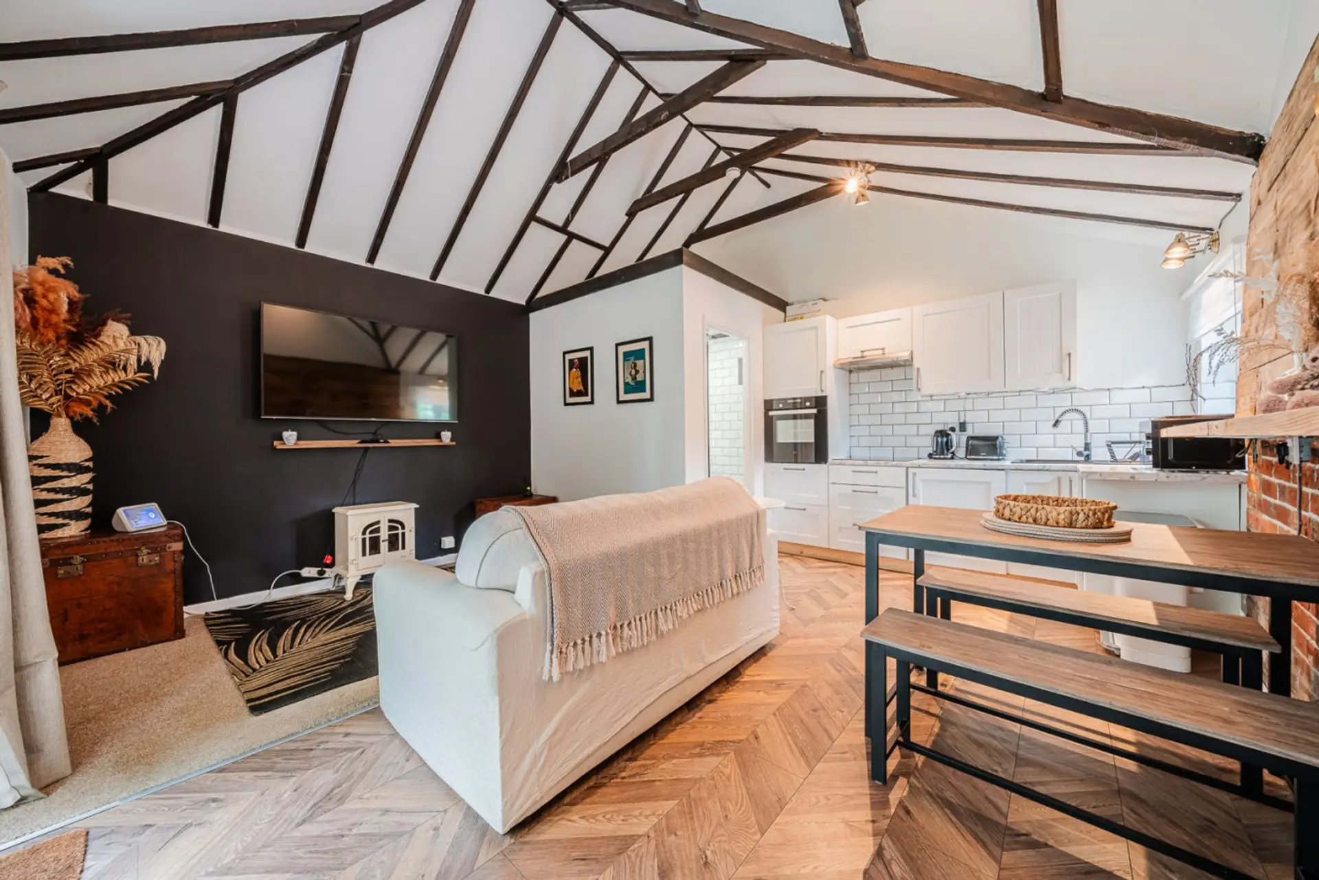 The open-plan living and dining area features a vaulted exposed-beam ceiling, white sofa, wooden bench dining table, and herringbone wood flooring.